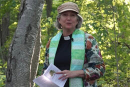 A female priest in a green stole leads liturgy in a forest